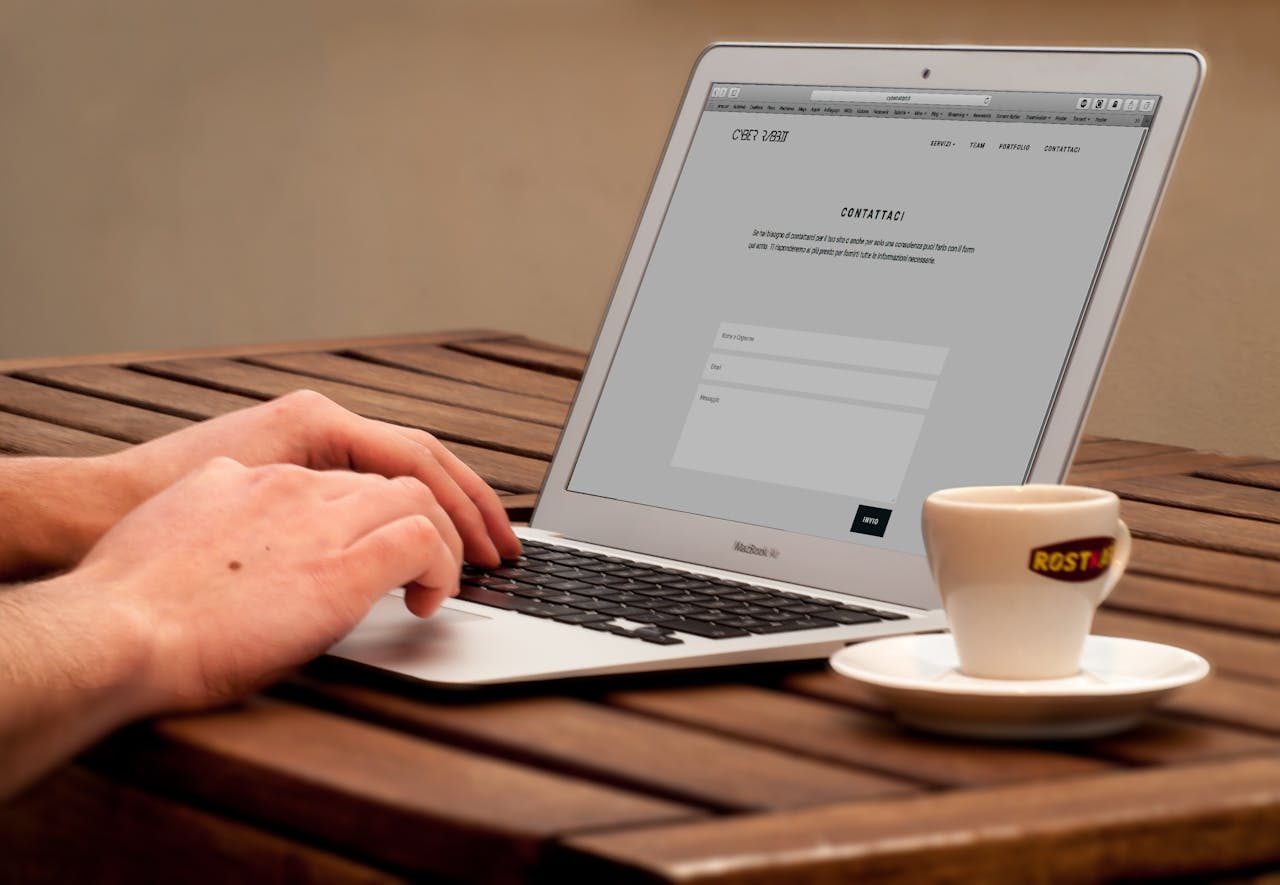 contact-header Close-up of hands typing on a laptop next to a cup of coffee on a wooden desk.